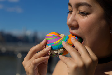 A beautiful young woman eats rainbow bagel with city in the background