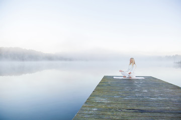 Woman relaxing and practicing yoga in the mist on the lake footbridge early morning. White clothes.