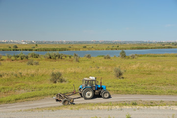 Трактор с сельскохозяйственным оборудованием едет по грунтовой дороге вдоль реки