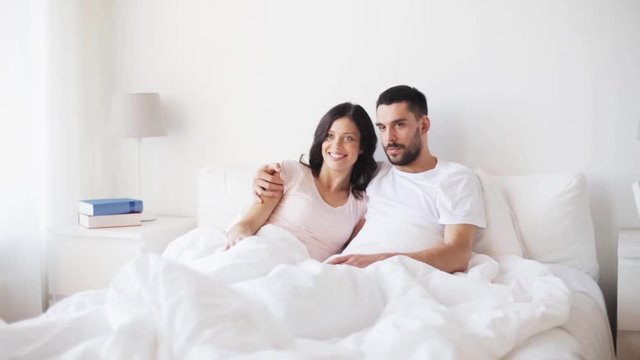 happy couple watching tv in bed at home