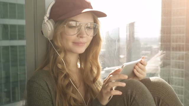 Pretty smiling girl sitting by window listening to music with headphones using mobile