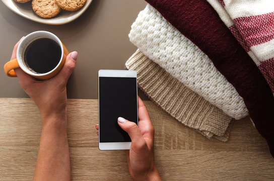 Hands holding coffee and phone on table with sweaters and cookies in autumn time