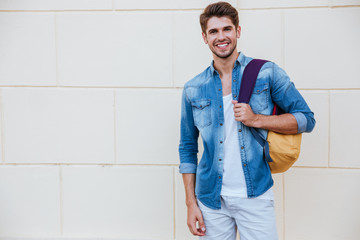 Cheerful young man with backpack standing and smiling - obrazy, fototapety, plakaty