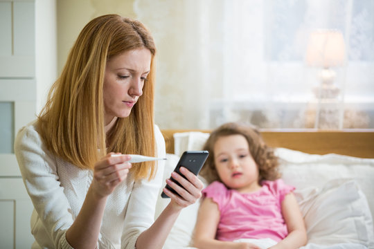 Mother Measuring Temperature Of Her Ill Kid. Sick Child With High Fever Laying In Bed And Mother Holding Thermometer. Mother With Cell Phone Calling To Doctor