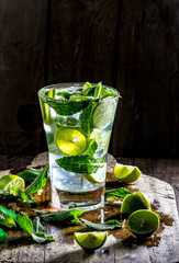 Glass of alcohol cocktail or lemonade. Mohito cocktail with lemon, lime, mint, ise cubes. Cold drink. Fresh lime and mint on wooden background