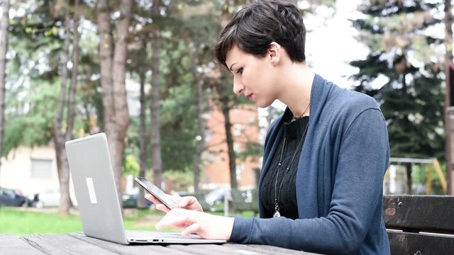 Yung beautiful caucasian brown hair woman sitting on a bench in a aprk using computer and smart phone &ndash; technology, multitasking, business concept