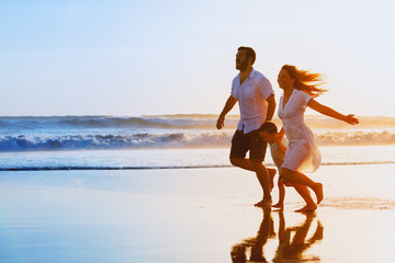 Happy family - father, mother, baby son hold hands and run together with fun along sunset sea surf on black sand beach. Travel, active lifestyle, parents with children on tropical summer vacations.