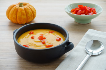 Pumpkin soup with chopped red peppers in a brown ceramic bowl with handles, a  munchkin pumpkin, a bowl with red chopped peppers, a grey napkin and a soup spoon on light brown wooden background.