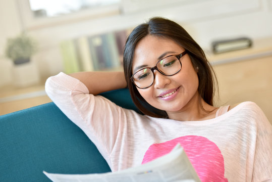 Asian girl with eyeglasses reading newspaper in armchair