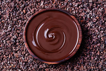 Bowl of melted chocolate on a crushed raw cocoa beans, nibs background. Copy space Top view