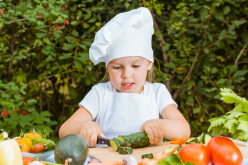 Девочка в костюме поваренка нарезает огурец на разделочной доске, лежащей на столе среди овощей