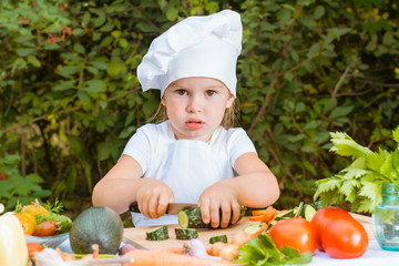 Девочка-поваренок нарезает огурец на разделочной доске, лежащей на столе среди овощей