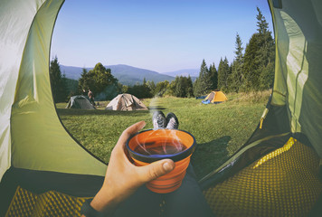 Woman lying in a tent with coffee ,view of camping with  people near the fire   and forest