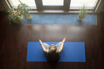 Attractive young woman working out at home, doing yoga exercise on blue mat, sitting in Easy Decent, Pleasant Posture , meditating, breathing, relaxing with closed eyes. Full length. Top view