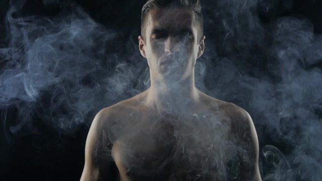 Handsome shirtless man with closed eyes standing in dark, with smoke around him.