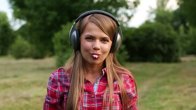 Young attractive girl with headphones bursts the bubble of chewing gum. Girl listening to music in a summer park.
