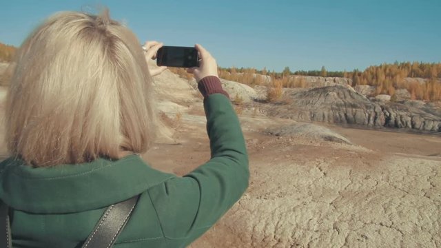 Young woman in a green coat taking pictures in the desert scenery with her smart phone admiring the landscape