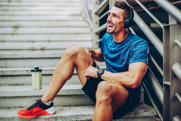 Handsome male jogger listening to music while taking break after morning training in urban setting