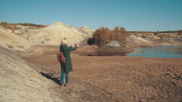 Young woman in a green coat taking pictures in the desert scenery with her smart phone admiring the landscape