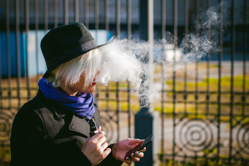 portrait of young beautiful woman with white hair, in a black coat, a skirt and a black hat, smoking an electronic cigarette, blowing the smoke vapor
