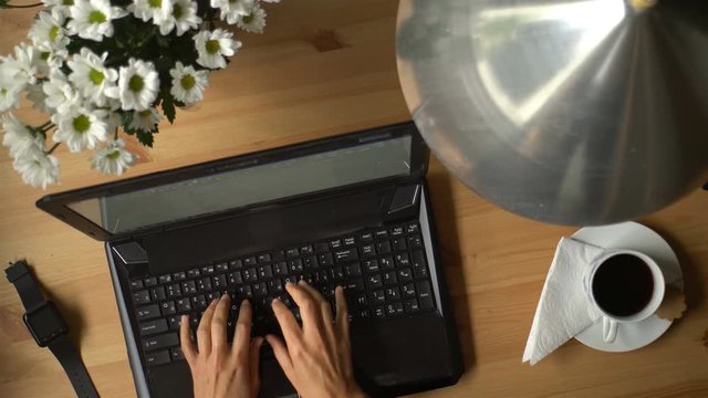 
A woman working at a computer with coffee and bouquet of flowers on the table. Top view.