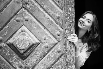 Pretty young girl peering from behind ancient wooden door, black-and-white photo.