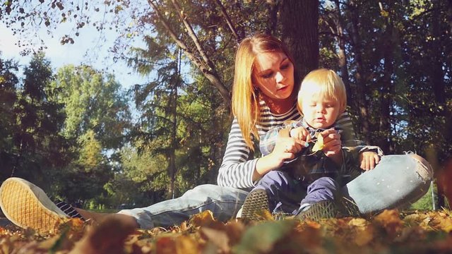 young beautiful mom playing with her baby. Mom and son sitting on the grass in a park in autumn in slowmotion. 1920x1080