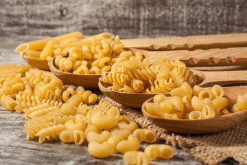 Various mix of pasta on wooden rustic background, sack and wooden spoons. Diet and food concept.