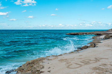 Beautiful view of rocky coast of azure coastline of Caribbean Sea in hot sunny summer day. Quiet small waves are splashing against coastal stones. Inscription Cuba on the sand. Clear serene blue sky