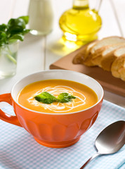 Pumpkin soup with cream and basil in orange bowl on a wooden table.