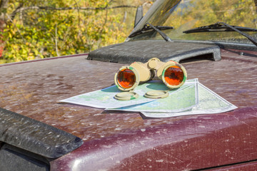 binoculars with a card lying on a dirty hood brutal off-road car on a background of yellow autumn foliage. journey orienteering locality find attractions navigitsii