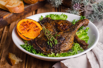 Confit duck legs with thyme and slice of orange in white plate on wooden background.  New year food. Selective focus