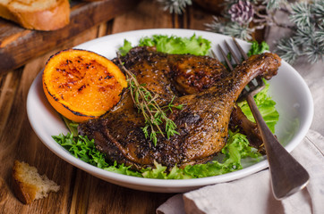 Confit duck legs with thyme and slice of orange in white plate on wooden background.  New year food. Selective focus
