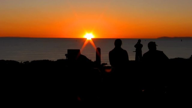 A couple sip wine, smoke cigars on a bench overlooking the ocean with a bicyclist taking pictures at sunset. HD 1080.