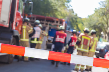 Fototapeta premium Team of blured firefighters by firetruck on protected accident site. Focus on red and white security band.