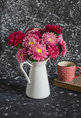 Bouquet of autumn flowers asters in a white jug, book and a mug of tea on a dark background. Still life in vintage style