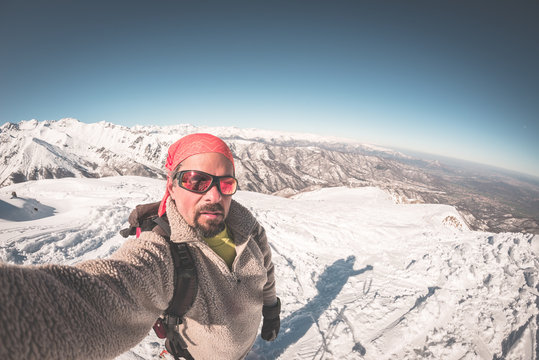 Adult alpin skier with beard, sunglasses and hat, taking selfie on snowy slope in the beautiful italian Alps with clear blue sky. Toned image, vintage style, ultrawide angle fisheye lens.