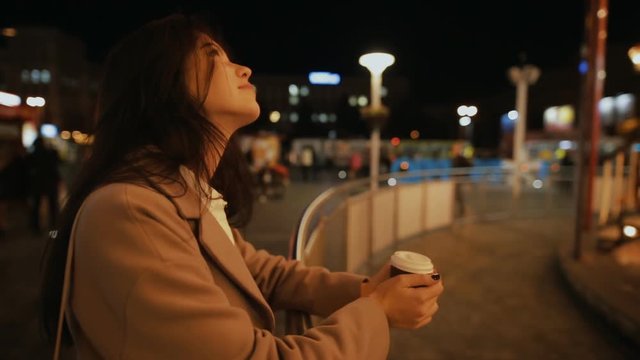 Woman looking to illuminated carousel in night city and drinking aroma coffee