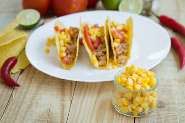 Yellow sweet corn in a small glass bowl and mexican tacos with chicken meat, chili pepper, tomato and corn as background