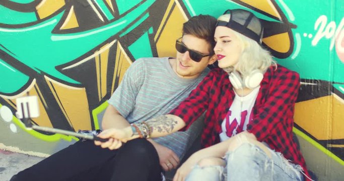 Trendy young hipster couple taking a selfie using a mobile on a stick as they pose together in the summer sun in front of colorful graffiti.