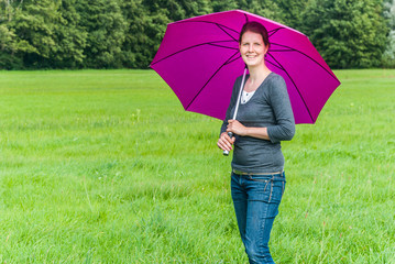 Young woman with brown hair smiling, looking at camera., holding a purple umbrella, standing on a meadow