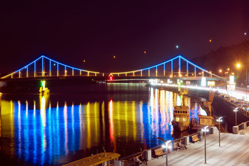 Night landscape. The city of Kiev, Ukraine, Europe. Pedestrian bridge across the Dnieper River. Beautiful lighting and reflection in water in blue and yellow colors