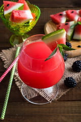 fresh watermelon in a glass with straws on a napkin. Triangular pieces of watermelon with mint and blackberries on a wooden board and a bowl. On a wooden background.