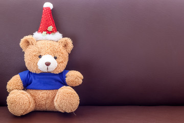 Brown teddy bear with blue shirt wearing christmas hat on the sofa
