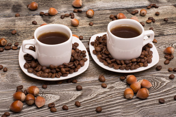 two small white cups of coffee with cocoa beans, slices of chocolate and hazelnuts on wooden background