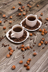 two small white cups of coffee with cocoa beans, slices of chocolate and hazelnuts on wooden background
