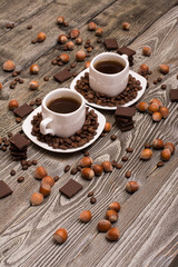 two small white cups of coffee with cocoa beans, slices of chocolate and hazelnuts on wooden background