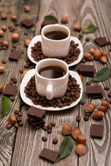 two small white cups of coffee with cocoa beans, slices of chocolate, hazelnuts and green leaves on wooden background