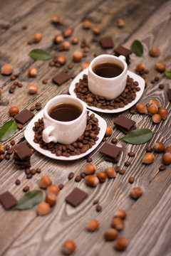 two small white cups of coffee with cocoa beans, slices of chocolate, hazelnuts and green leaves on wooden background