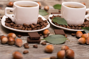 two small white cups of coffee with cocoa beans, slices of chocolate, hazelnuts and green leaves on wooden background